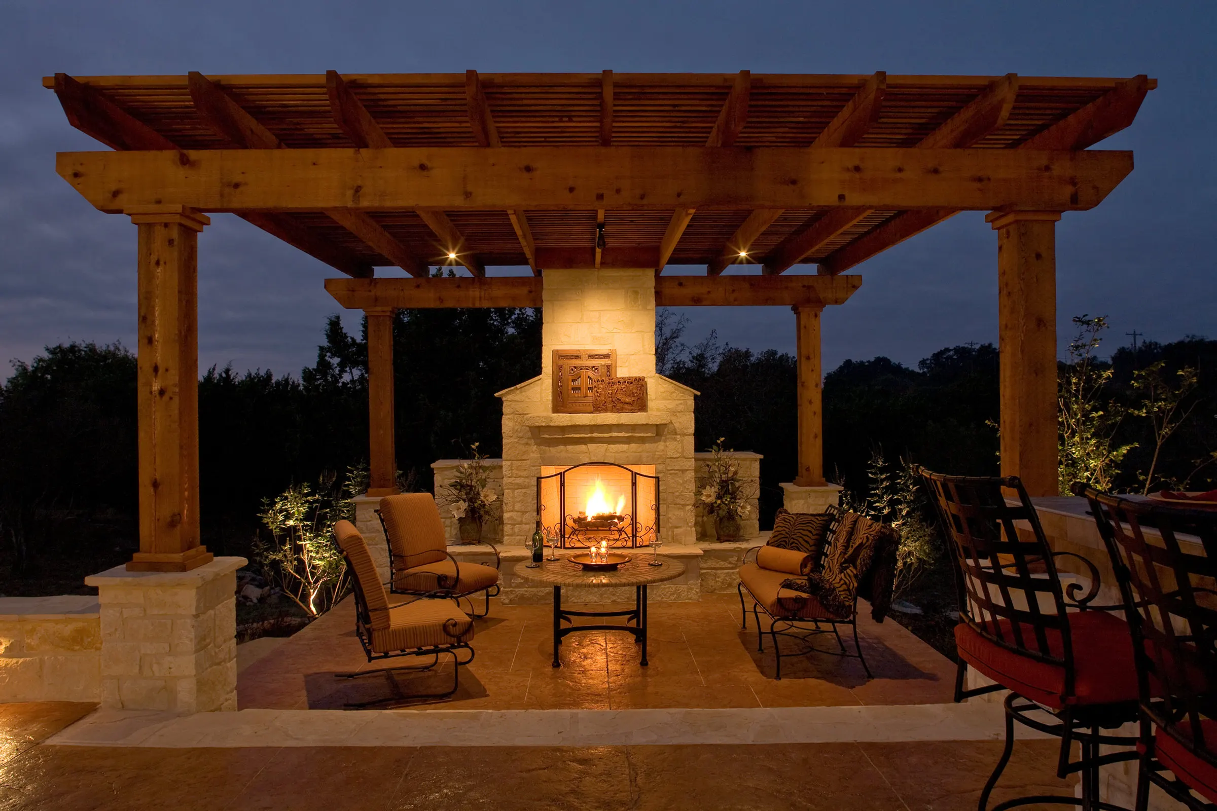 Stone outdoor fireplace and seating beneath a timber pergola at dusk
