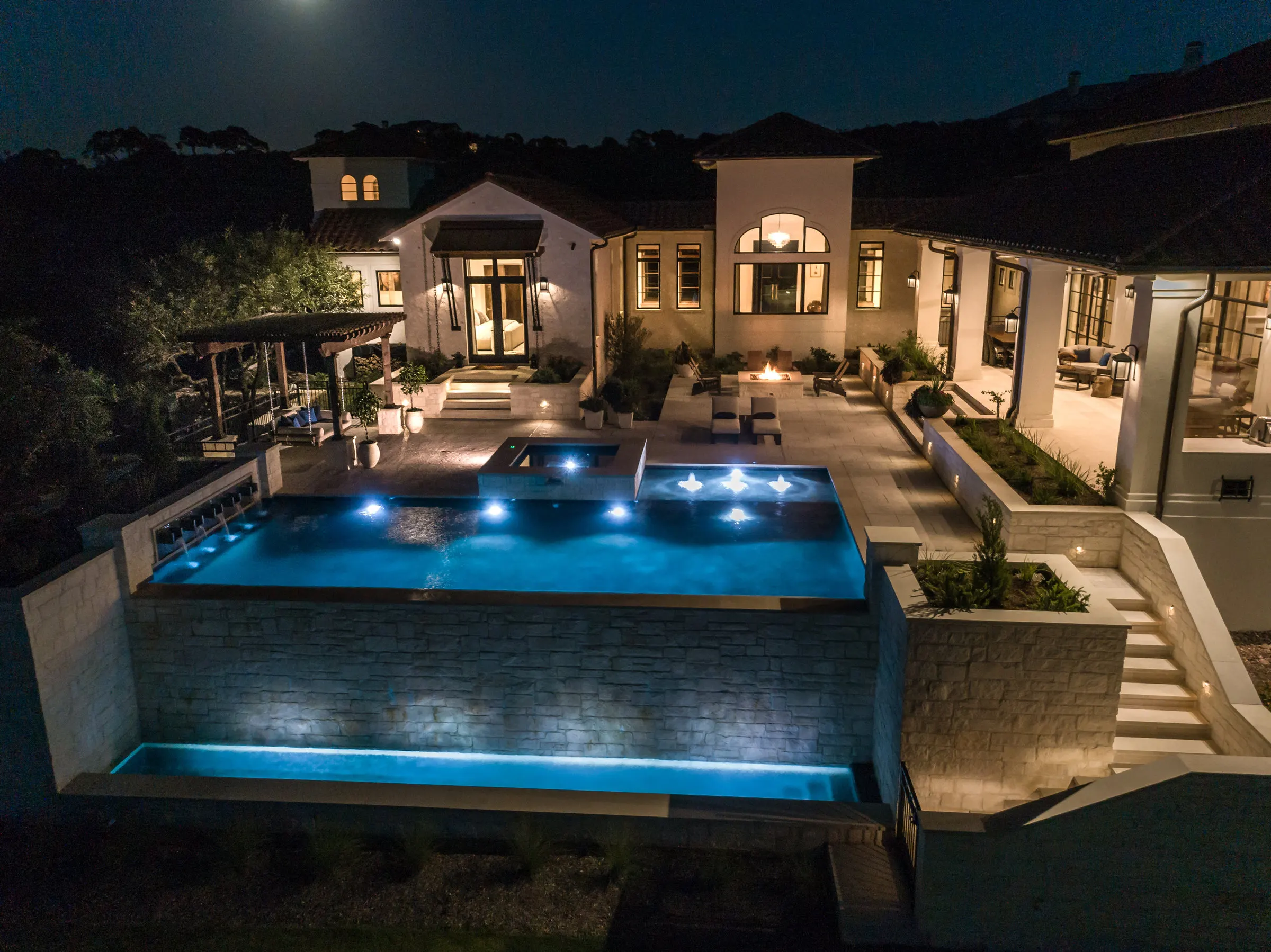 Contemporary vanishing edge pool lit at night, with a tall stone wall above the slope